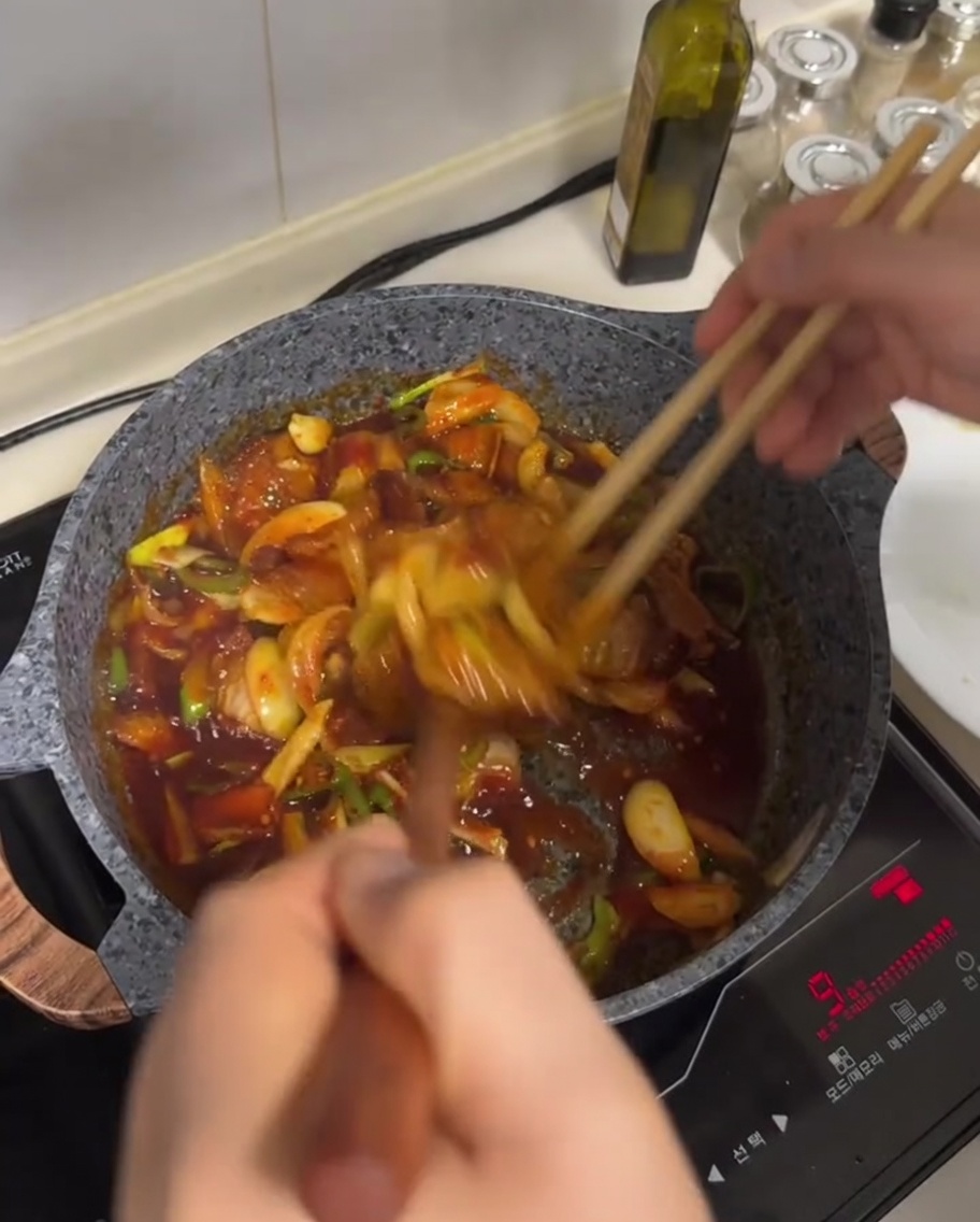 Salteado de panceta y verduras siendo mezclado con una salsa roja en una sartén.