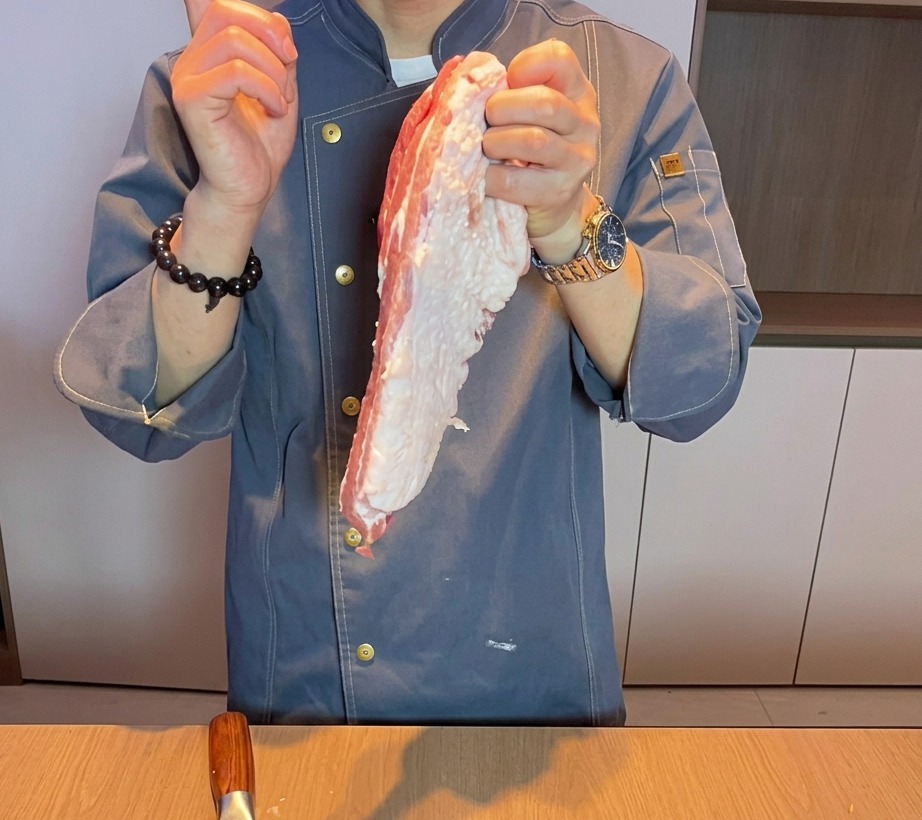 Primer plano de una mano de chef cortando panceta de cerdo fresca y cruda en trozos gruesos sobre una tabla de cortar de madera.