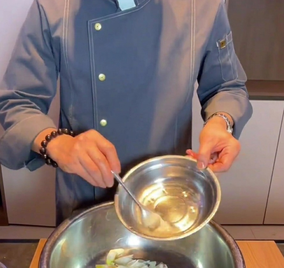 Mano vertiendo agua con bicarbonato en un bol de mezcla de metal con trozos de panceta de cerdo cruda, jengibre y rodajas de cebolleta.