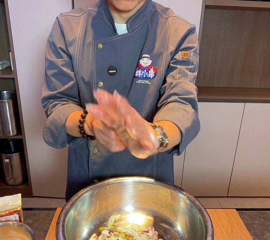 Chef con un guante transparente mezclando panceta de cerdo cruda y aromáticos en un bol grande de metal.