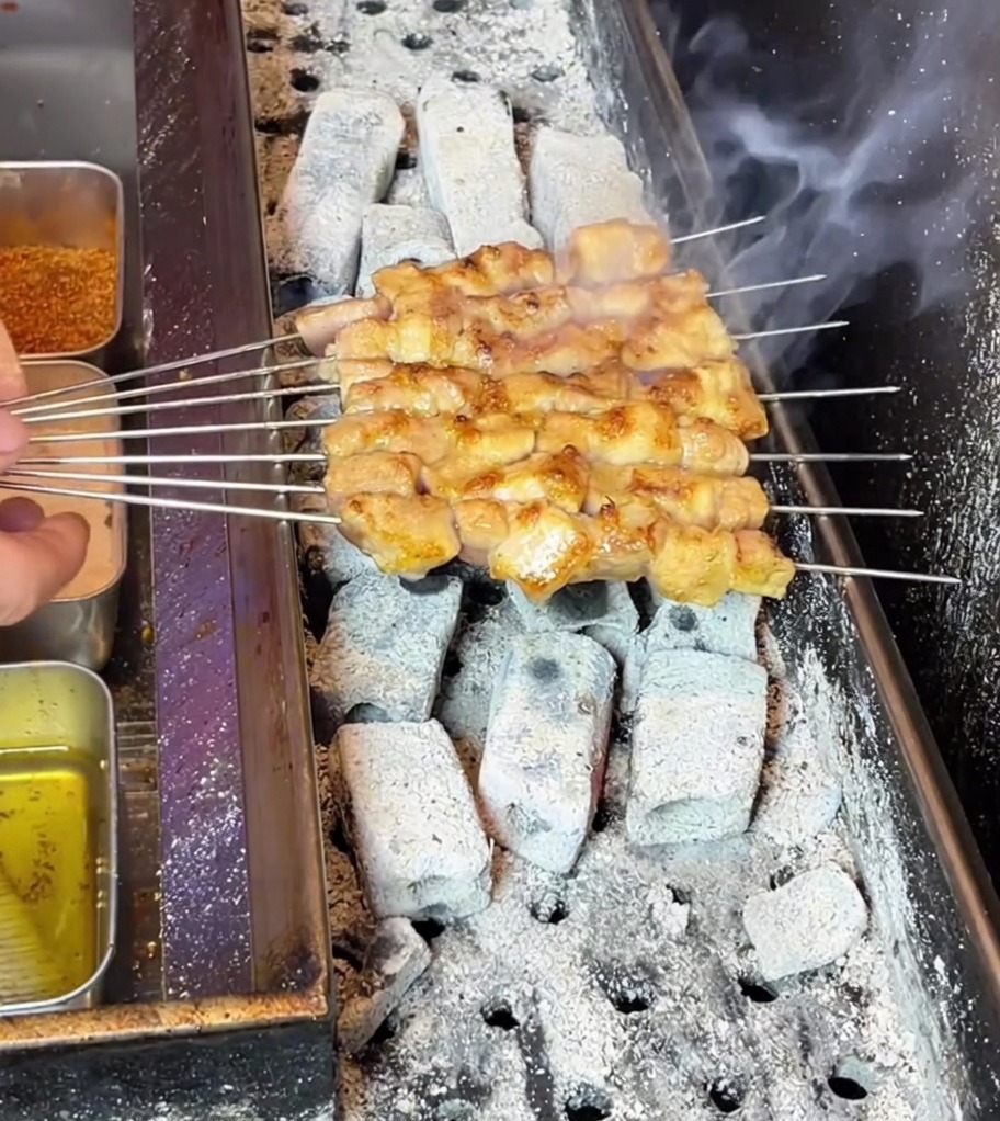 Brochetas de panceta de cerdo volviéndose doradas mientras chisporrotean sobre una parrilla de carbón caliente y ahumada.