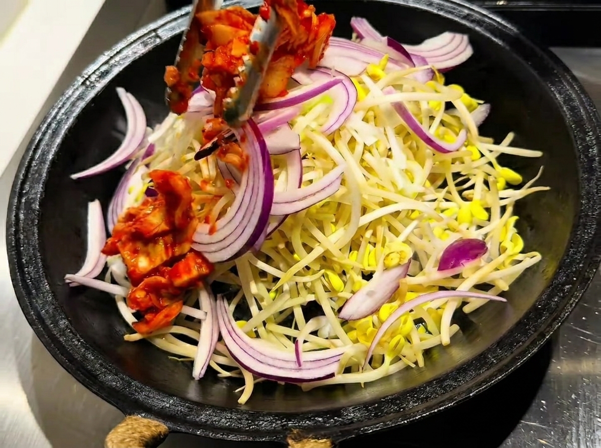 Una sartén que contiene una capa de brotes de soja cubierta con kimchi rojo vibrante y cebollas en rodajas.