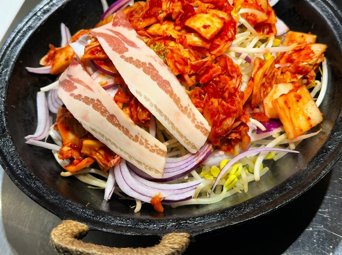 Rodajas de panceta de cerdo cruda dispuestas con cuidado sobre una capa de kimchi, cebollas y brotes de soja.