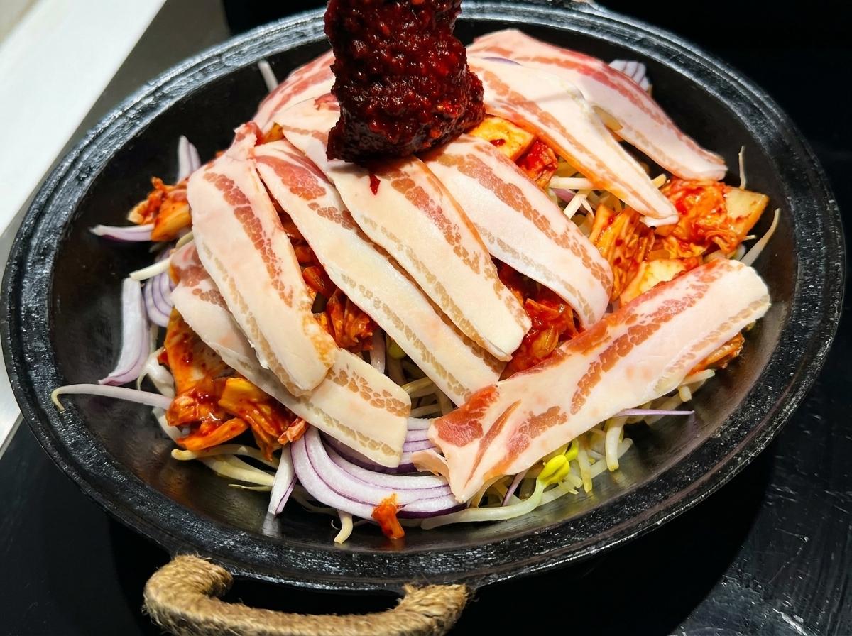 Salsa coreana roja vibrante para saltear siendo extendida sobre una sartén con panceta en capas y verduras frescas.