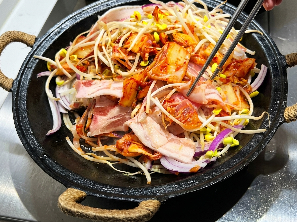 Pinzas mezclando panceta en rodajas, brotes de soja, kimchi y salsa en una olla de hierro fundido.