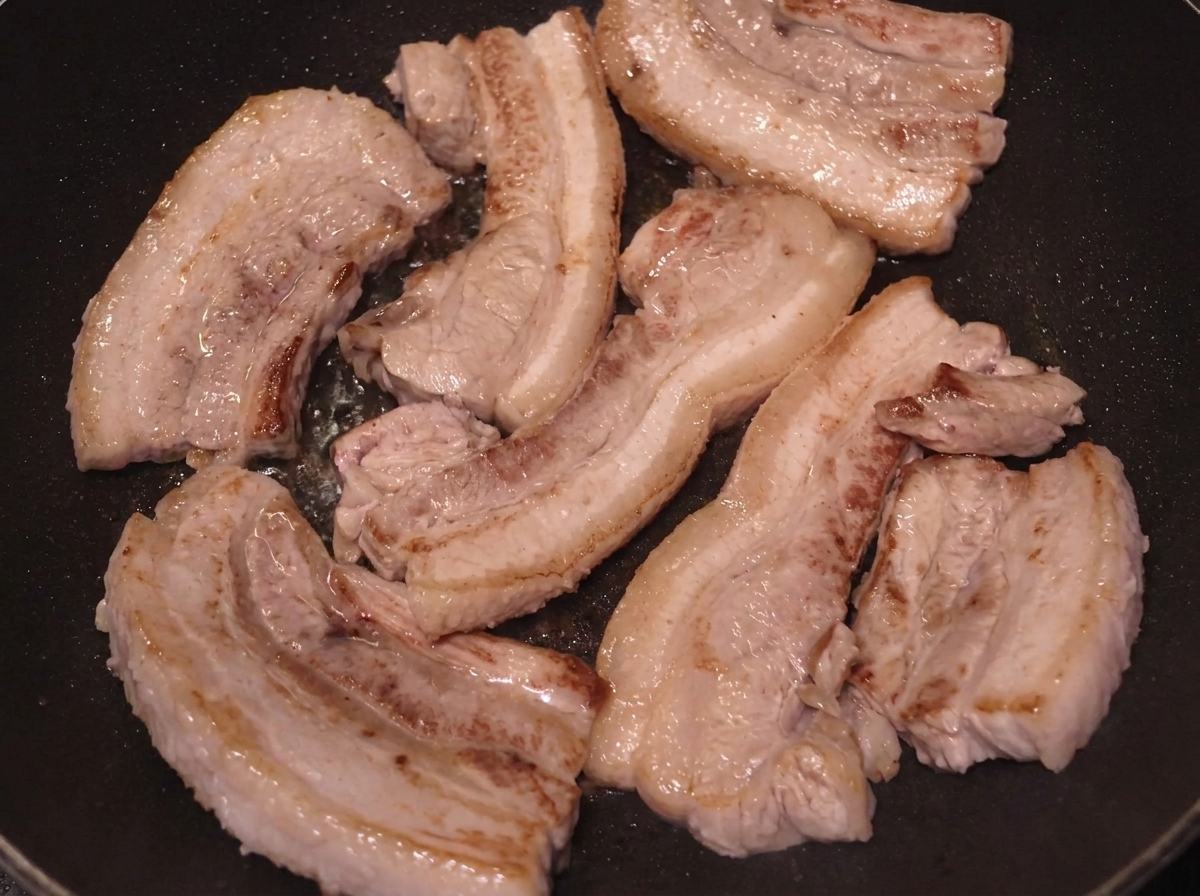 Varios trozos de panceta dorándose en una sartén con superficies bien marcadas.