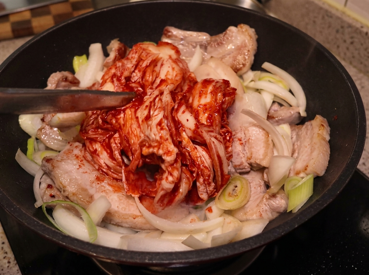Un trozo grande de kimchi entero colocado con pinzas sobre la panceta y cebollas en la sartén.