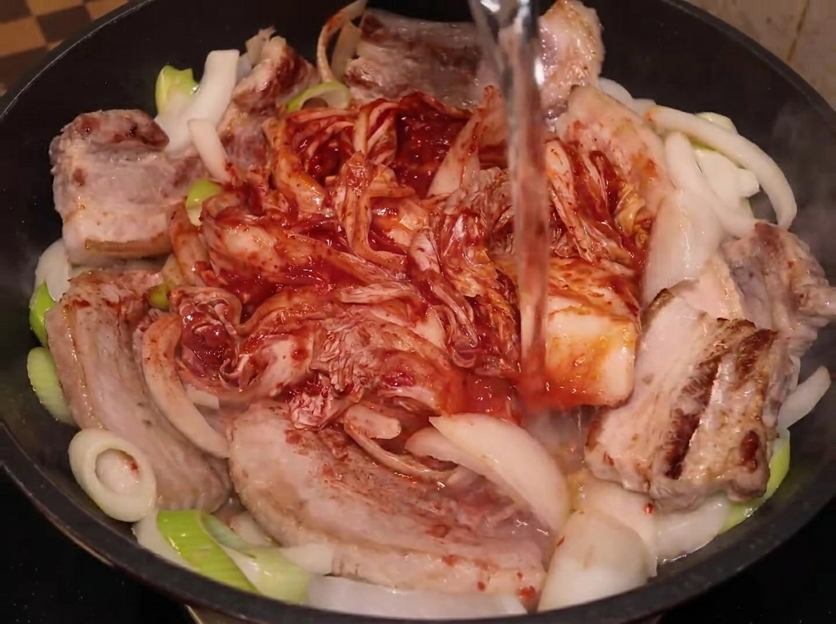 Agua siendo vertida desde una botella en una sartén con panceta, cebollas y el kimchi entero.