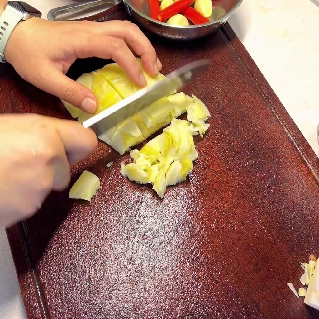 Manos sosteniendo un cuchillo y cortando trozos de col encurtida de color verde claro sobre una tabla de cortar de madera oscura.