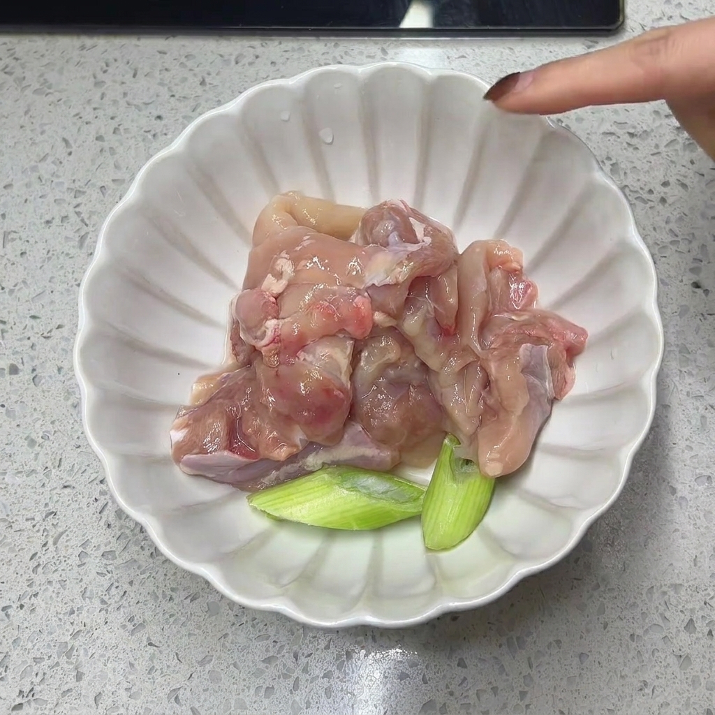 Carne de muslo de pollo cruda y deshuesada junto a trozos de cebolleta en un tazón blanco.