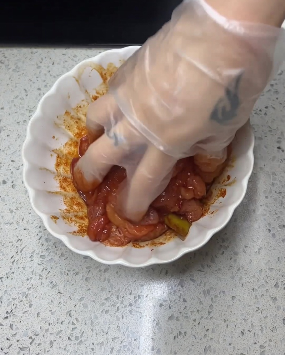 Una mano con guante masajeando una marinada roja y especiada sobre piezas de pollo crudo en un tazón blanco.