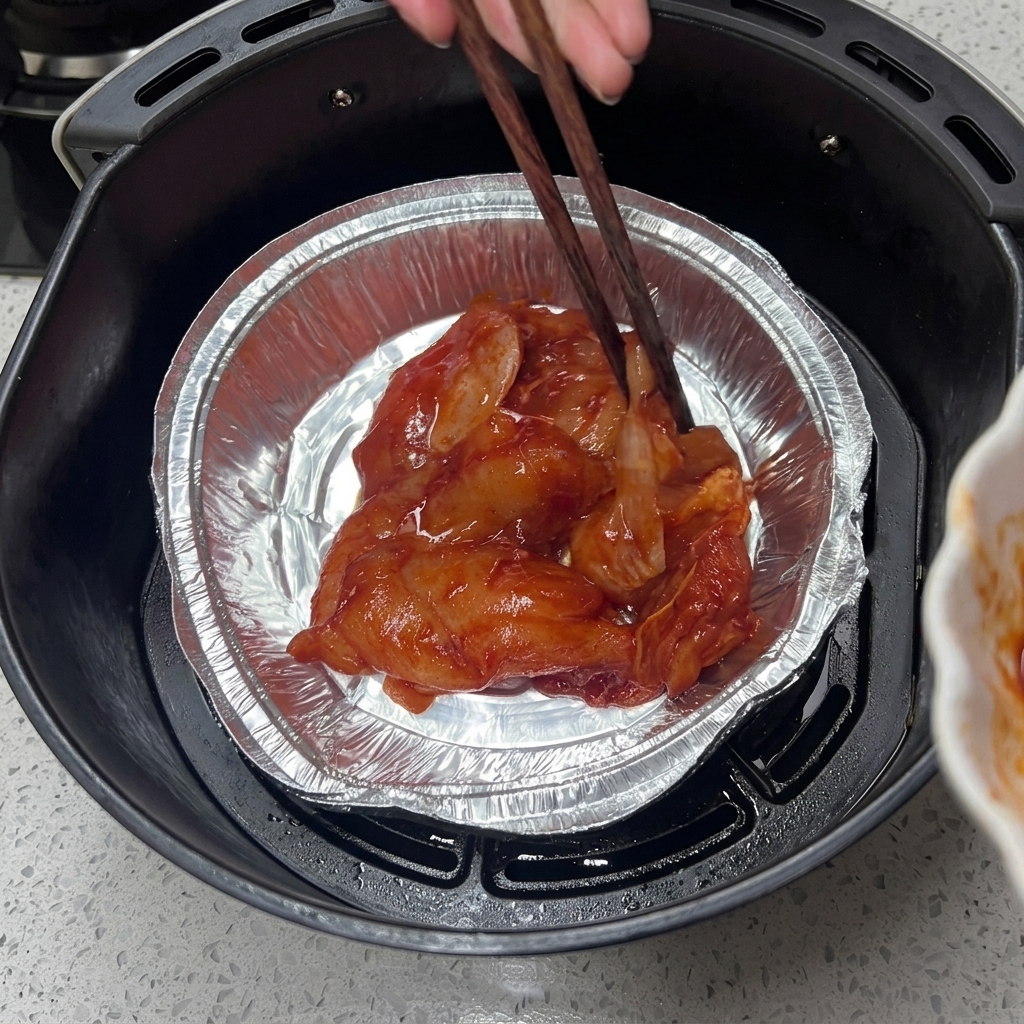 Palillos de madera acomodando piezas de pollo crudo marinado en una bandeja de papel de aluminio dentro de la canasta de la freidora de aire.