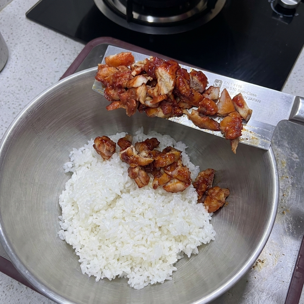 Pollo cocido picado deslizándose desde un cuchillo ancho hacia un tazón grande de metal lleno de arroz blanco.