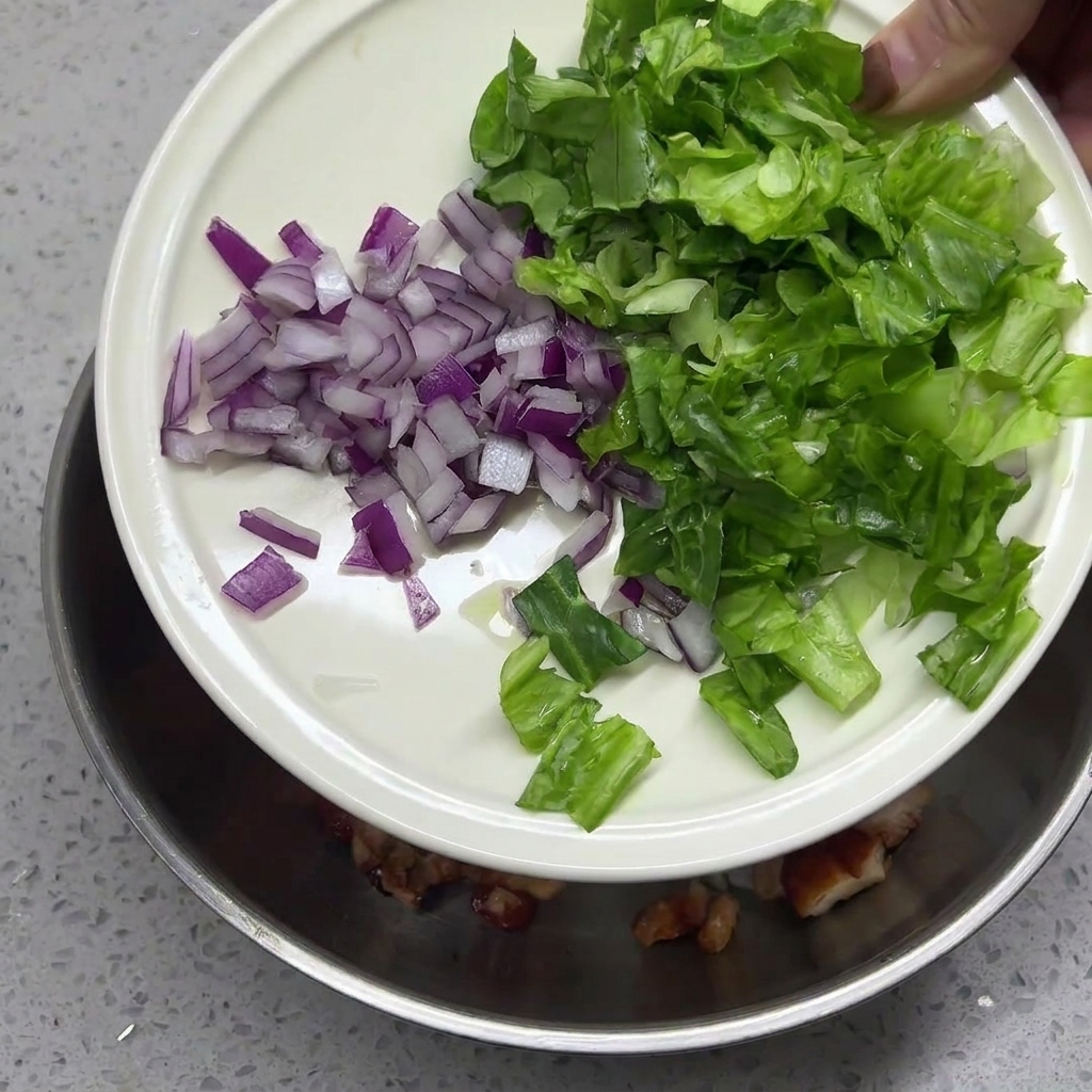 Cebolla roja picada y lechuga verde en tiras vertiéndose desde un plato blanco a un tazón que contiene pollo y arroz.