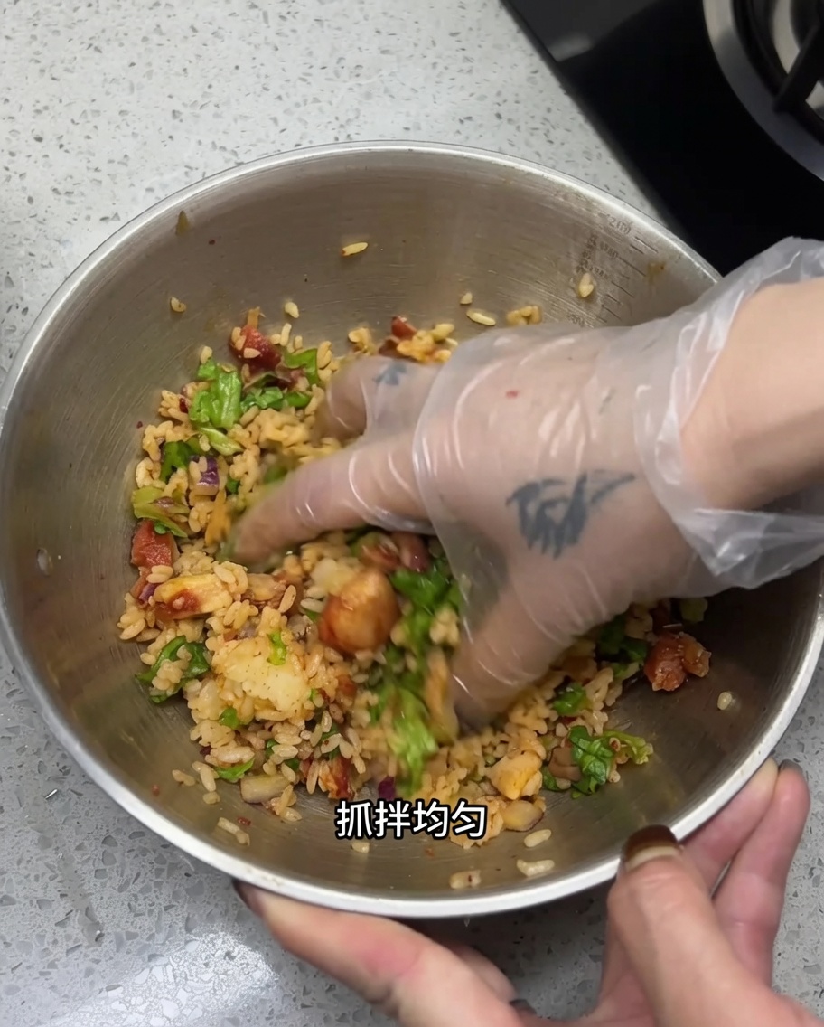 Una mano con guante mezclando el arroz, el pollo picado y las verduras en un tazón grande de acero inoxidable para asegurar que todo quede bien combinado.
