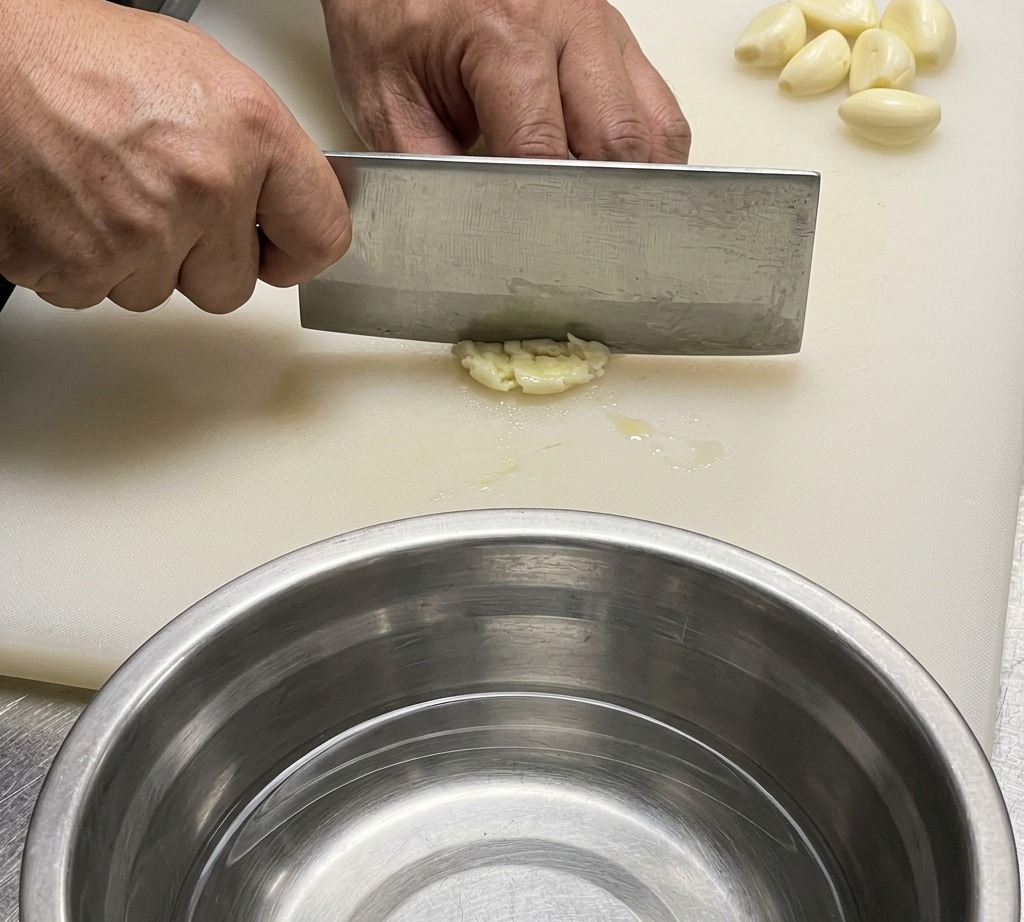 Un primer plano de un cuchillo de chef picando ajo y jengibre en una tabla de cortar blanca.