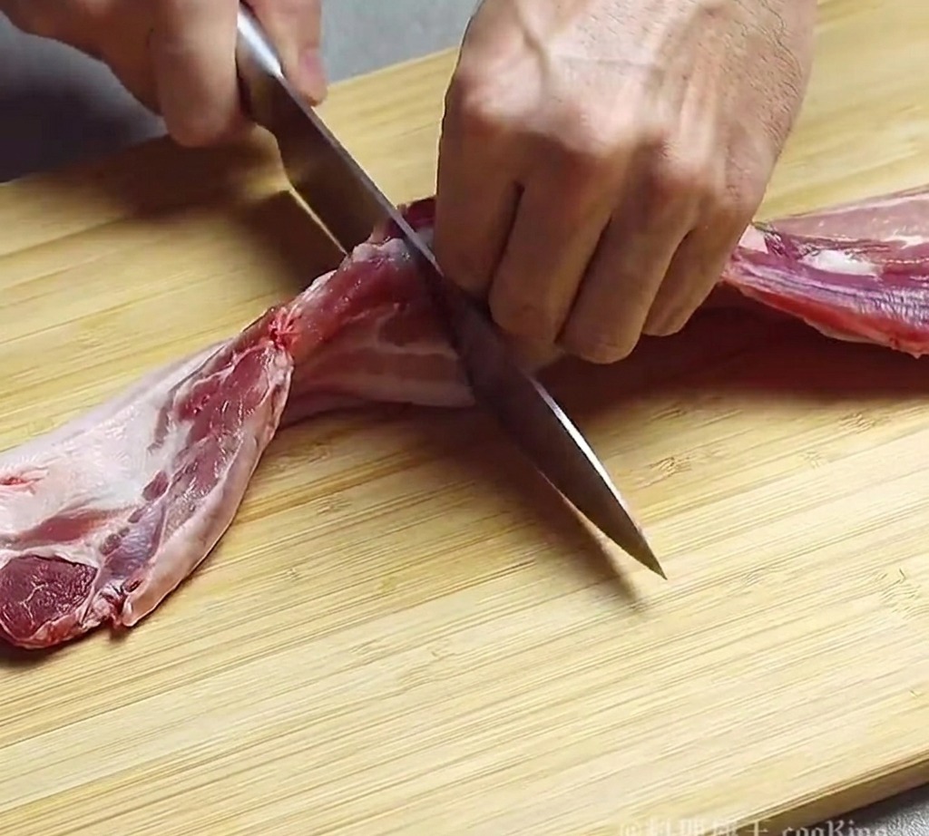 Una persona usando un cuchillo de chef para cortar un trozo de panceta cruda en tiras gruesas sobre una tabla de madera.