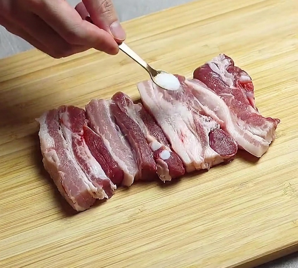 Tiras de panceta cruda dispuestas sobre una tabla de madera siendo sazonadas con una pizca de sal.
