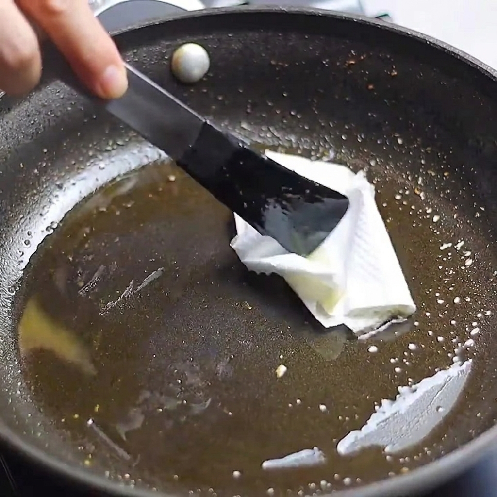 Un papel de cocina sujeto con pinzas siendo usado para limpiar el exceso de aceite de una sartén.