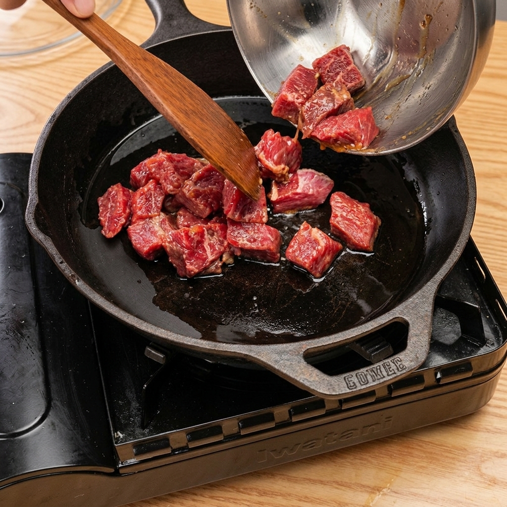 Cubos de ternera dorándose en una sartén de hierro fundido, con vapor subiendo tras añadir vino de cocina y tapar.