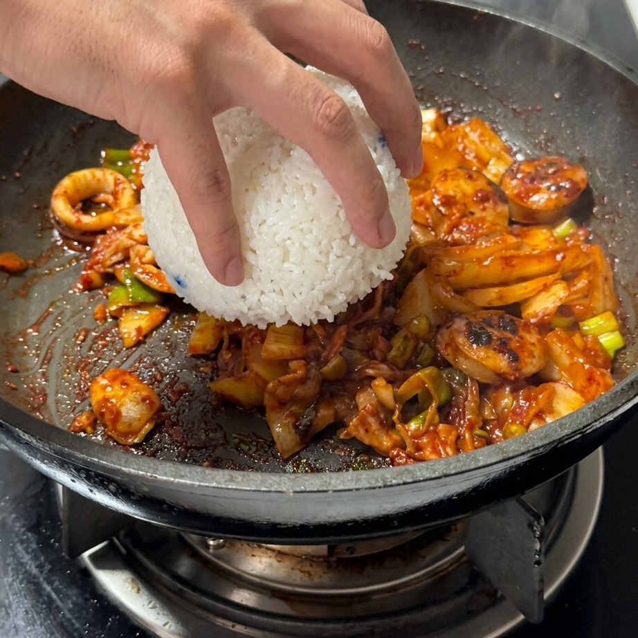 Un bol de arroz blanco al vapor siendo añadido a la sartén con la mezcla de kimchi y carne.