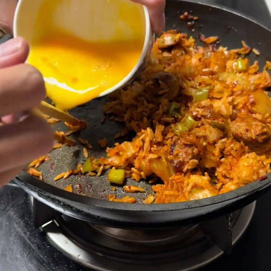 Huevo batido siendo vertido en un lado despejado de la sartén, junto a la mezcla de arroz frito.