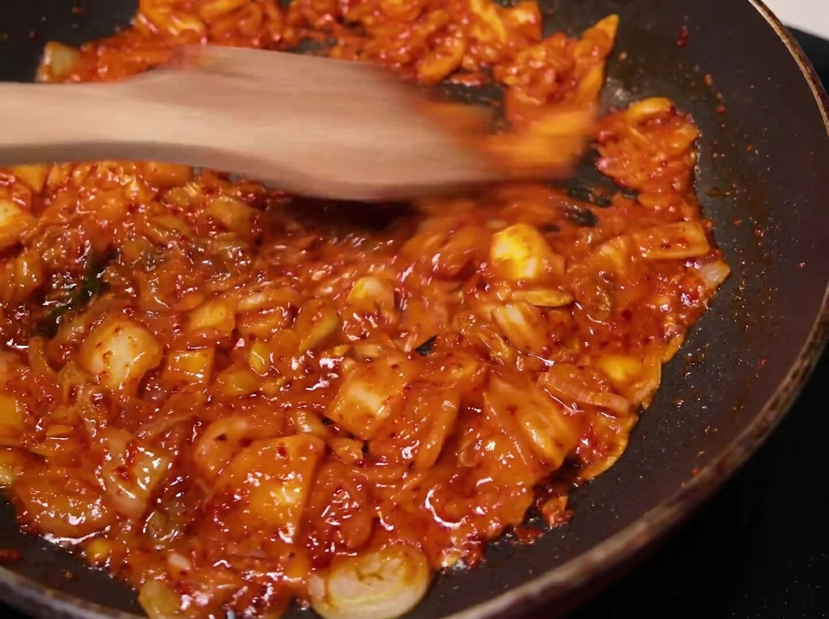 La mezcla de kimchi sazonado siendo removida y cocinada en una sartén con una espátula de madera.