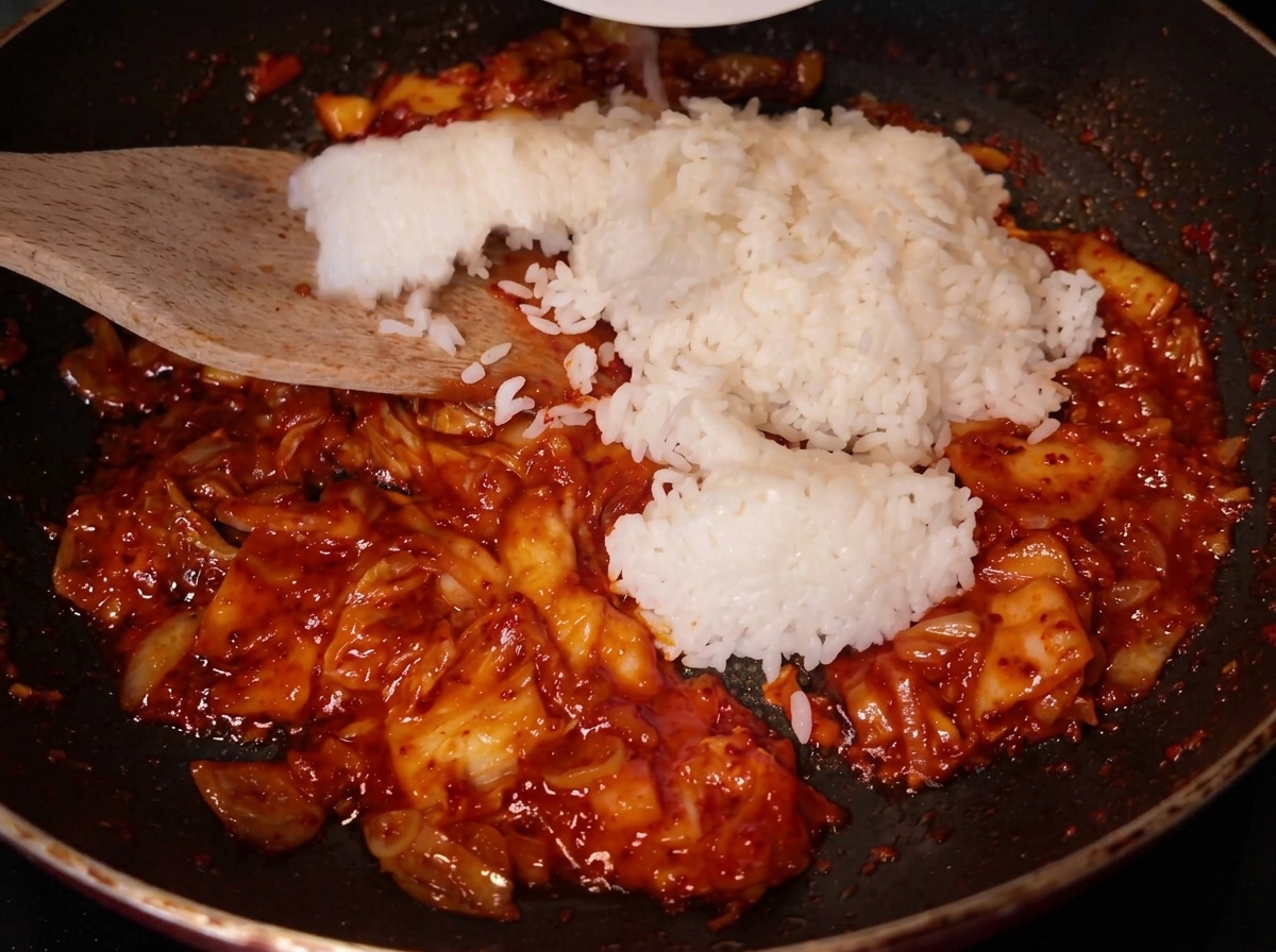 Una espátula de madera añadiendo una gran porción de arroz blanco en la sartén donde ya se está salteando la mezcla roja de kimchi.