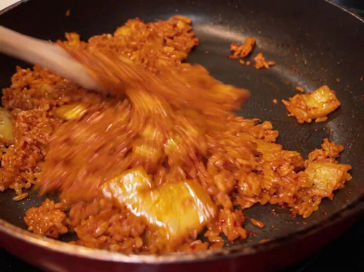 Un primer plano del arroz siendo mezclado con el kimchi rojo en la sartén, mostrando cómo el arroz adquiere un color rojizo-anaranjado uniforme.