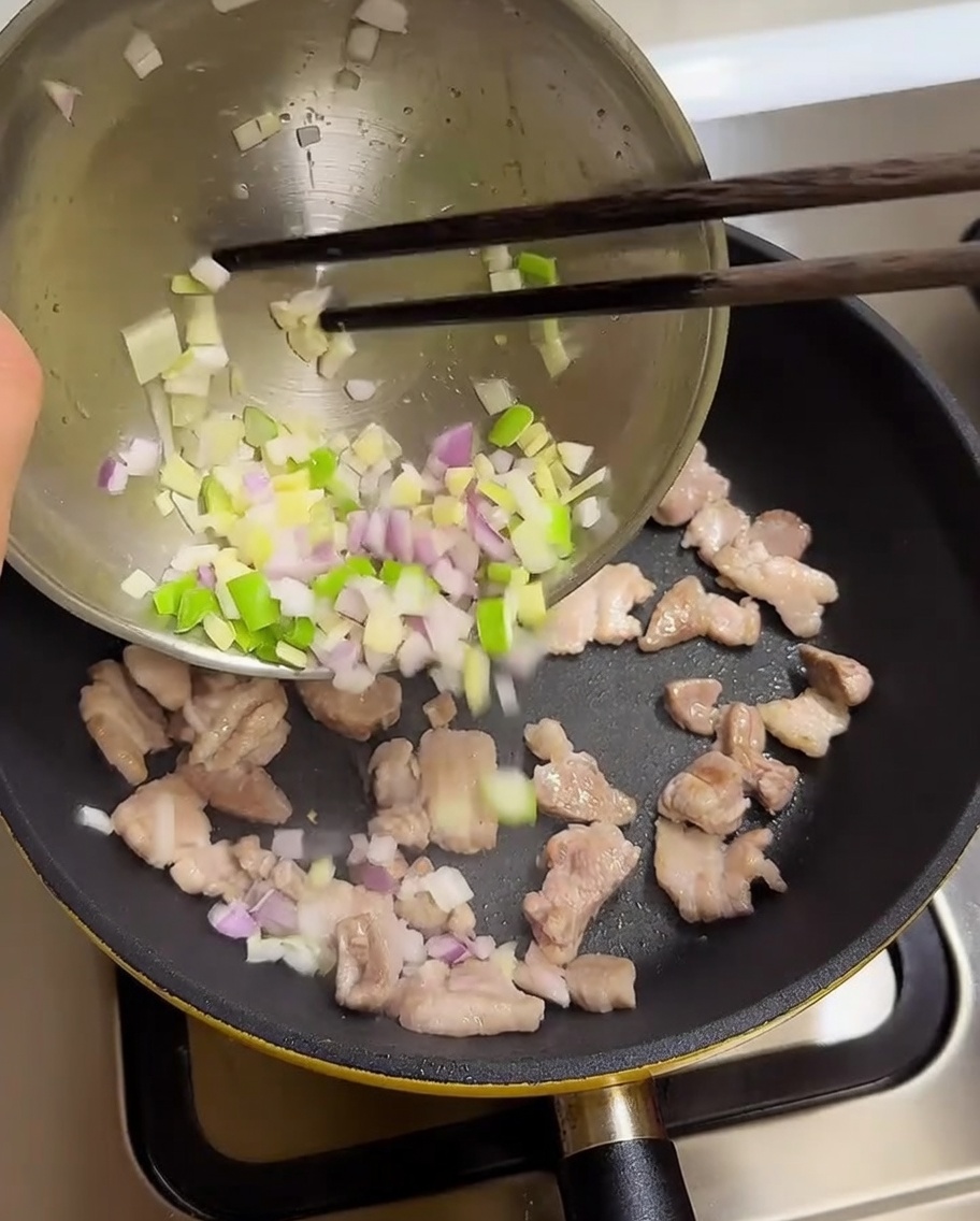 Cebolletas, ajo y cebolla picados vertiéndose desde un bol a una sartén con panceta de cerdo cocinada.