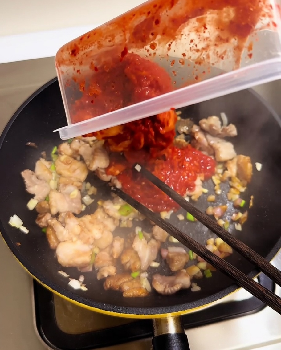 Kimchi rojo picado vertiéndose desde un recipiente a una sartén con cerdo y aromáticos.