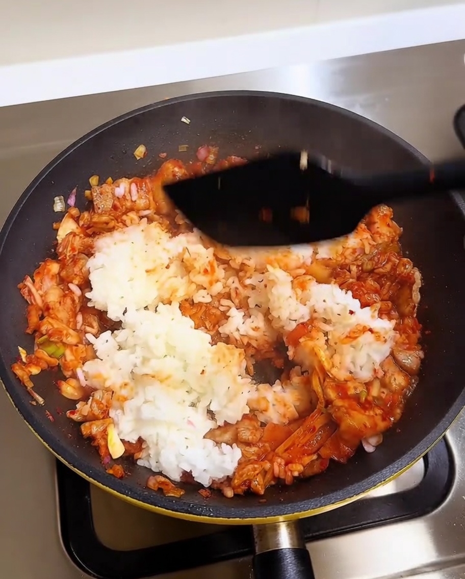 Una espátula mezclando arroz blanco con una vibrante base roja de cerdo y kimchi en una sartén caliente.