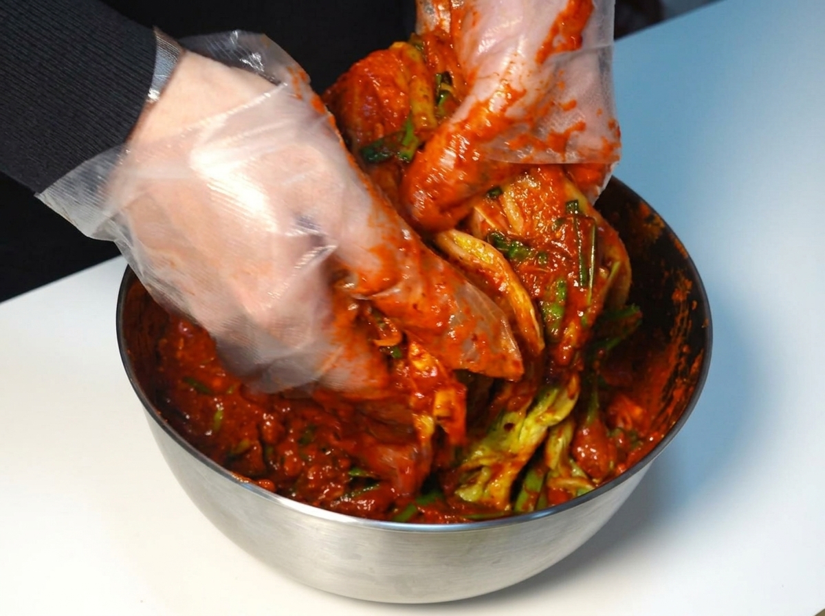 Manos enguantadas extendiendo pasta roja sobre cada capa de la col napa.