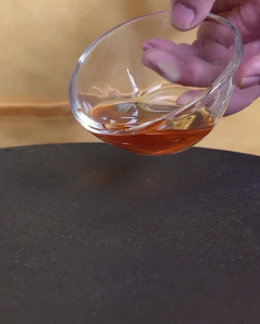 Un primer plano de aceite de sésamo vertiéndose desde un cuenco de cristal en una sartén caliente vacía.