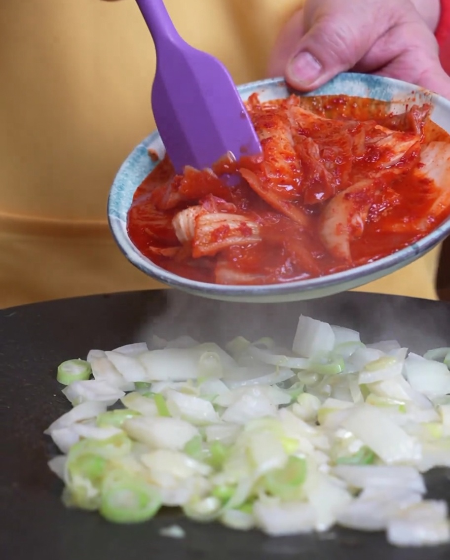 Kimchi siendo añadido a la sartén donde ya se están cocinando las cebolletas.