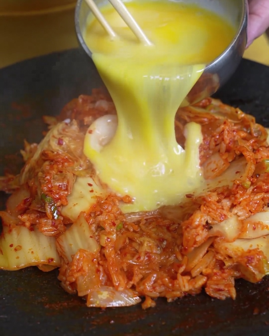 Huevos batidos siendo vertidos sobre una montaña de arroz frito con kimchi en una sartén caliente.