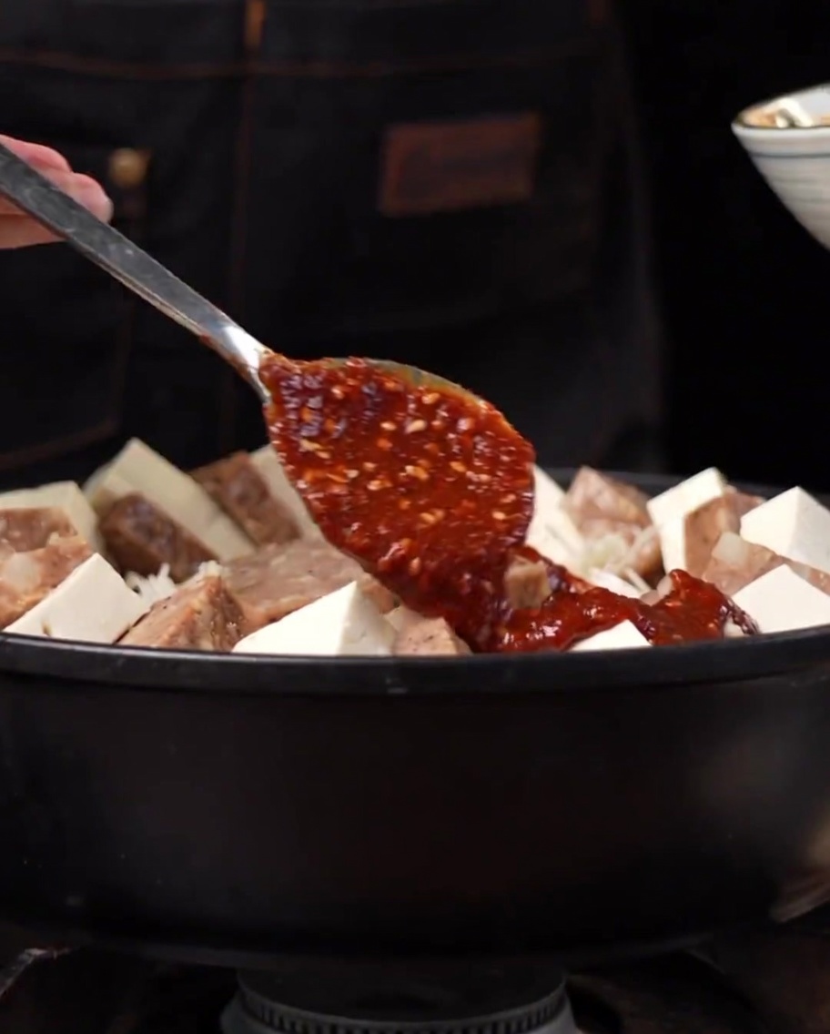 Una salsa picante rojiza siendo extendida sobre los ingredientes dispuestos en una olla de metal.