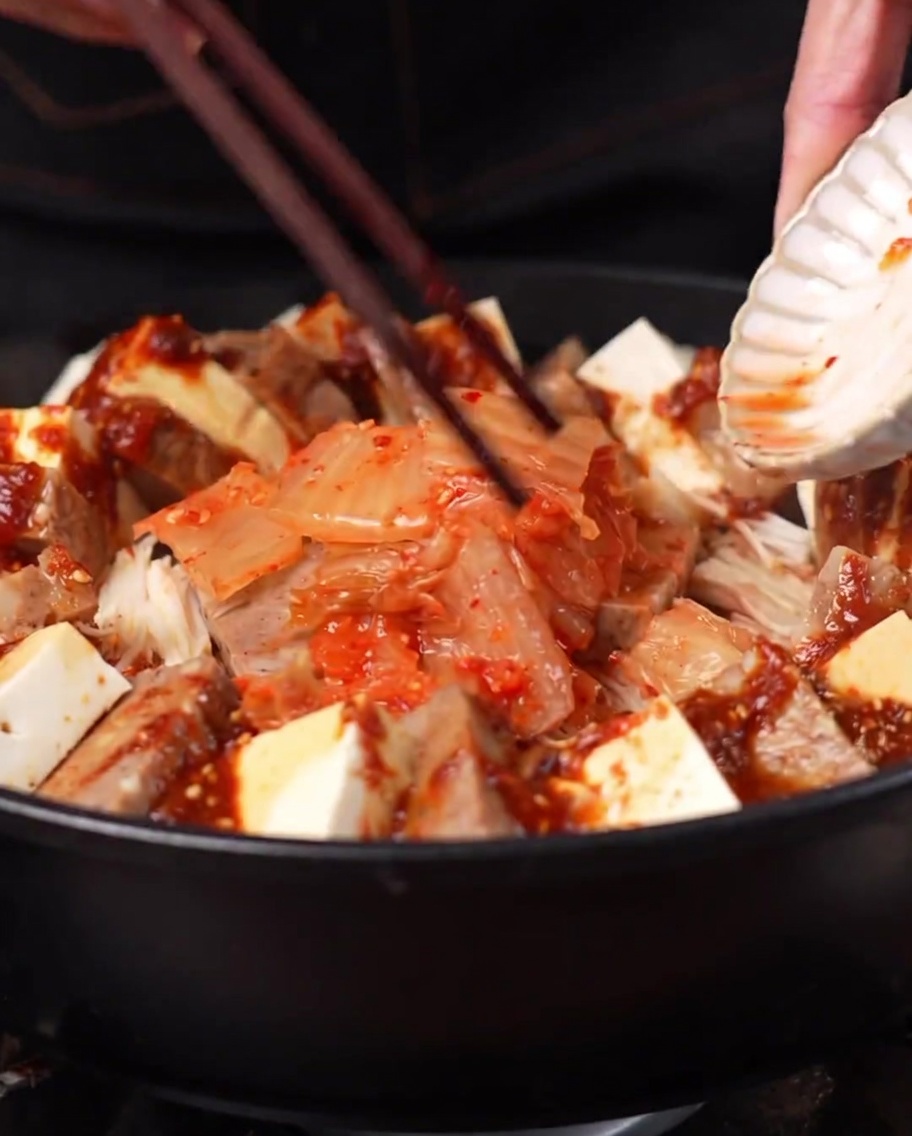 Una porción de kimchi siendo añadida al centro de la olla rodeada por los otros ingredientes.