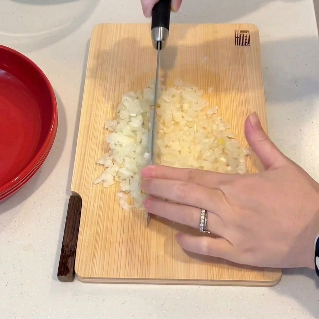 Una mano usando un cuchillo de chef para picar finamente una cebolla sobre una tabla de cortar de madera.