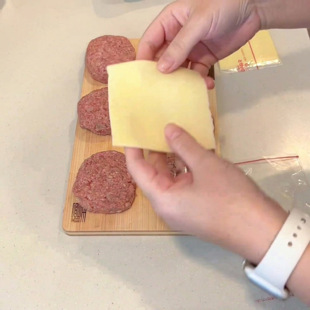 Una loncha de queso doblada siendo colocada en el centro de una hamburguesa de carne cruda aplanada.