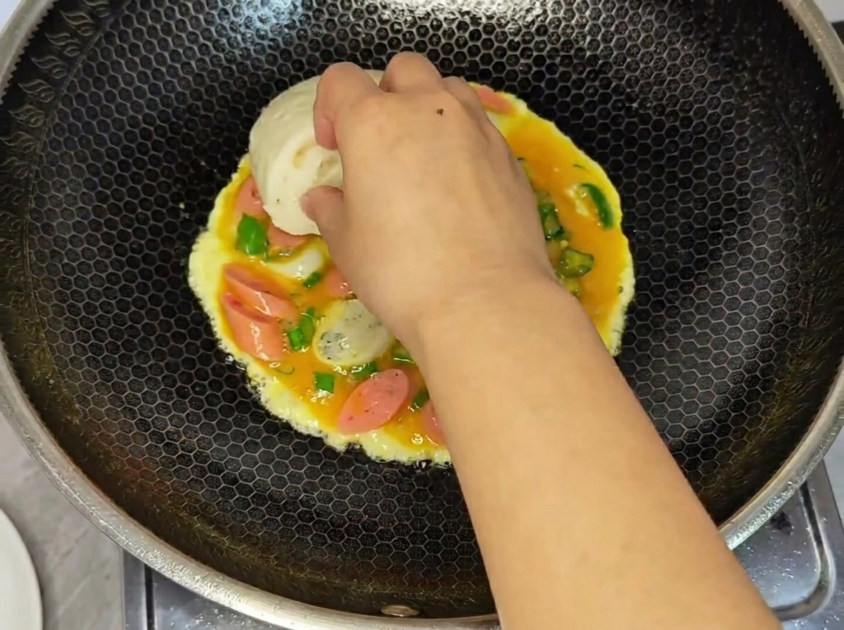 Una mano presionando una mitad de un pan al vapor blanco sobre la mezcla húmeda de huevo y salchicha cocinándose en un wok.