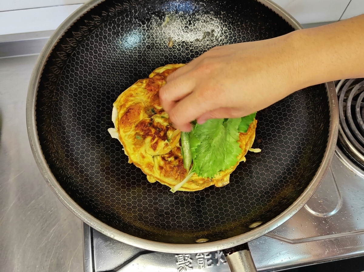 Una mano colocando una hoja de lechuga fresca sobre la tortilla de huevo cocida en un wok.