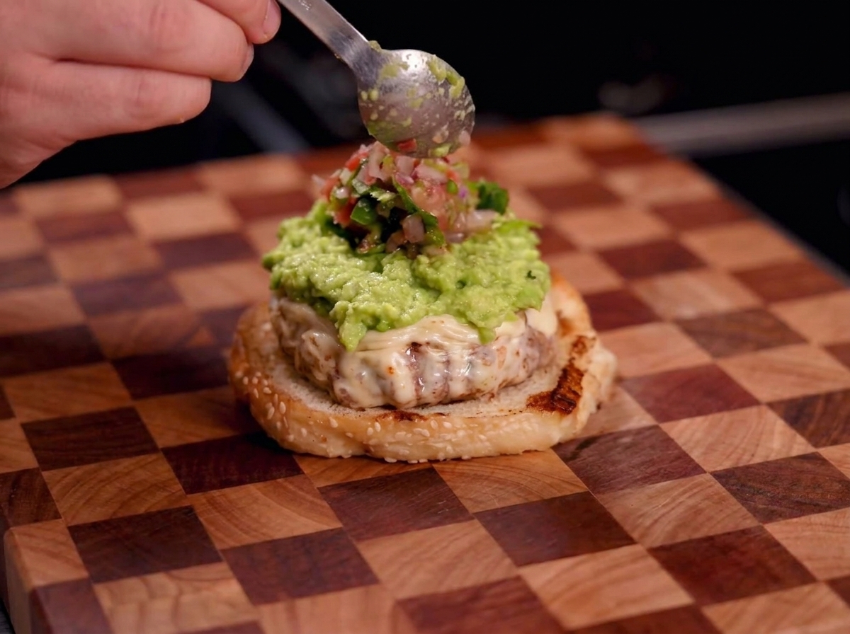 Una mano usando una cuchara para poner salsa roja fresca sobre la capa de guacamole verde en una hamburguesa.