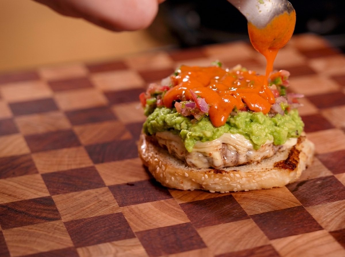 Salsa picante de color naranja brillante cayendo de una cuchara sobre una capa de salsa fresca en una hamburguesa.