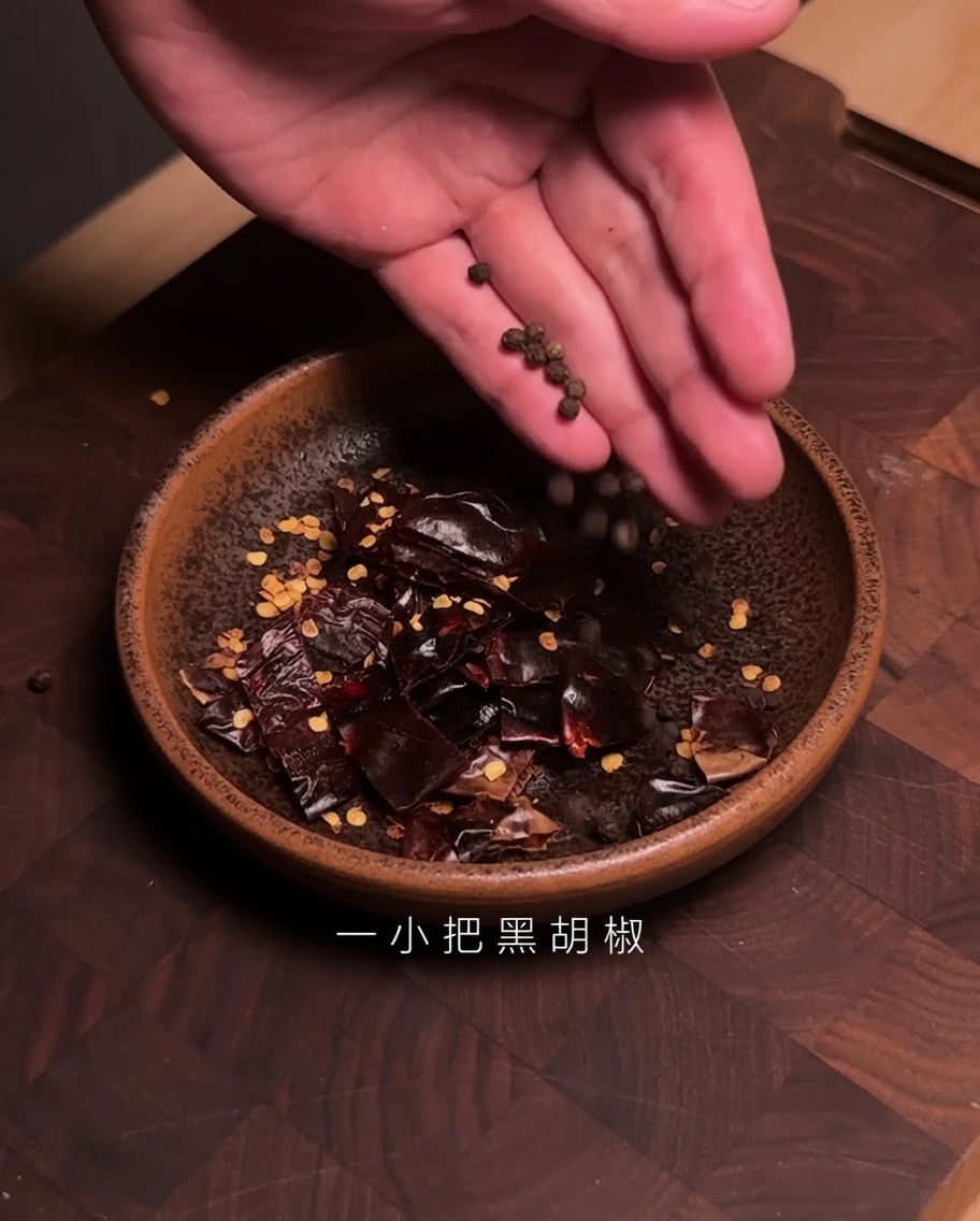 Manos dejando caer granos de pimienta negra en un tazón que contiene trozos de chile Guajillo cortado.