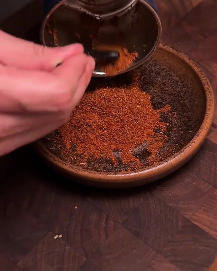 Un molinillo de especias vertiendo chile rojo recién molido y especias en un tazón de cerámica marrón.