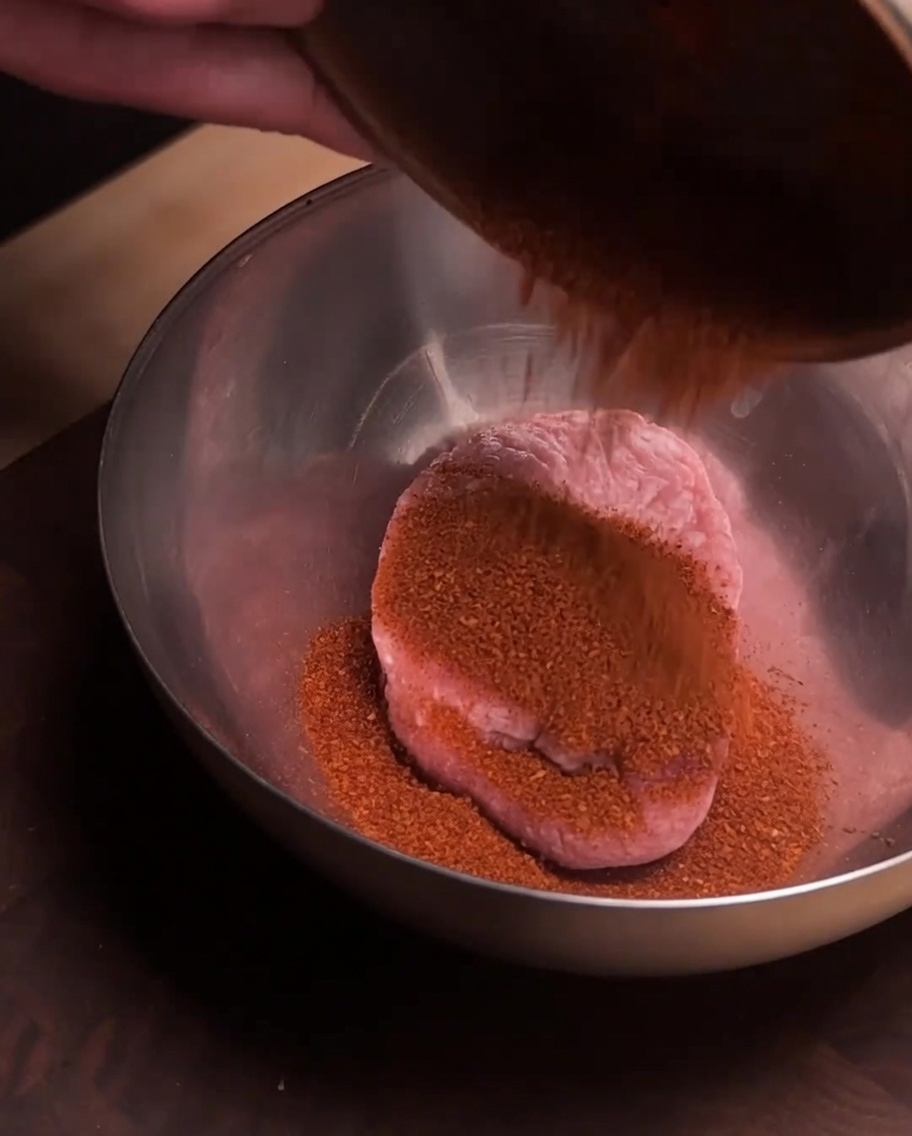 Carne de cerdo molida fresca en un tazón de acero inoxidable con una capa de polvo de especias rojo encima.