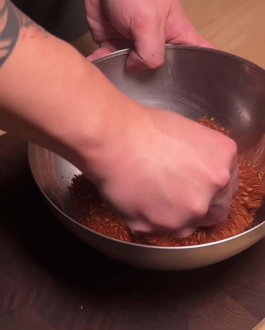 Una mano mezclando la carne de cerdo sazonada y el polvo de especias rojo en un tazón de acero inoxidable.