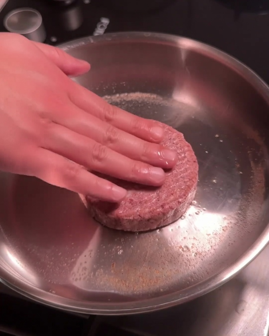 Una hamburguesa de res cruda siendo presionada hacia abajo en una sartén caliente de acero inoxidable.