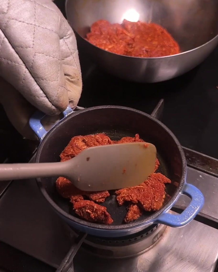 Una pequeña olla azul en la estufa con carne de cerdo molida siendo removida con una espátula de color claro.